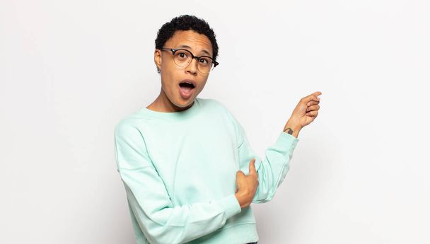 young afro woman feeling shocked and surprised, pointing to copy space on the side with amazed, open-mouthed look - 写真・画像