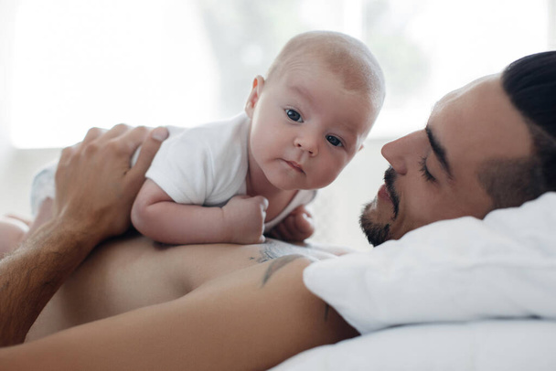 man with infant. happy family at home. happy father with cute two months baby boy laying in light bed at home - Photo, Image
