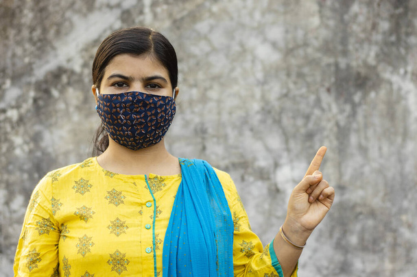 an Indian woman wearing homemade cotton nose mask, looking at camera and pointing left side for corona virus or covid-19 protection - Zdjęcie, obraz