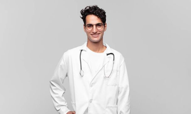 νέος γιατρός χαμογελά ευτυχισμένος με το χέρι στο ισχίο και αυτοπεποίθηση, θετική, υπερήφανη και φιλική στάση - Φωτογραφία, εικόνα