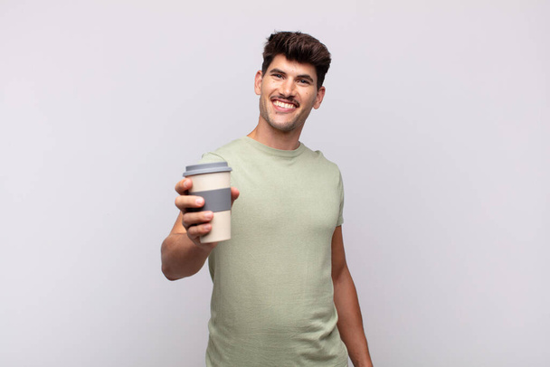 young man with a coffee smiling happily with a hand on hip and confident, positive, proud and friendly attitude - Фото, зображення