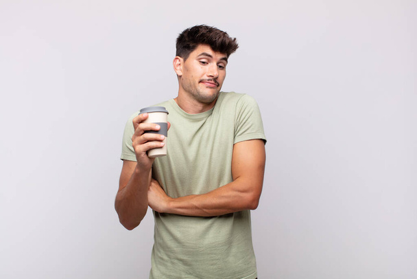 young man with a coffee shrugging, feeling confused and uncertain, doubting with arms crossed and puzzled look - Foto, imagen