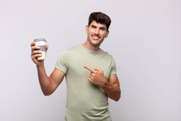 young man with a coffee smiling cheerfully, feeling happy and pointing to the side and upwards, showing object in copy space - Fotografie, Obrázek