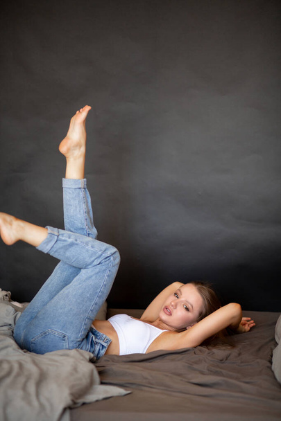 Beautiful girl in jeans and a top is lying on the bed with her legs up - Фото, изображение