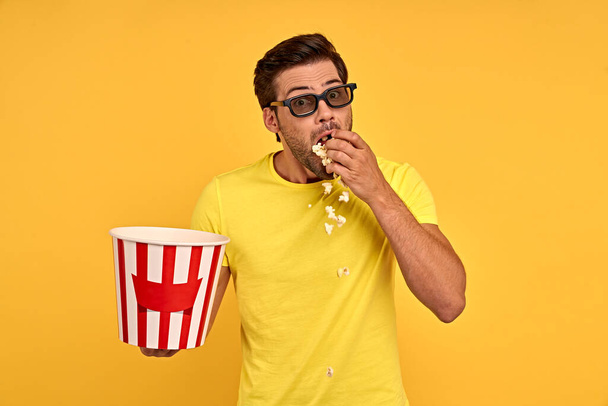 Schockierter, verängstigter junger Mann in lässiger Kleidung, 3D-Brille auf gelbem Hintergrund. Menschen Emotionen im Kino, Lifestylekonzept. Film ansehen, Eimer mit Popcorn halten und Mund mit der Hand bedecken. - Foto, Bild