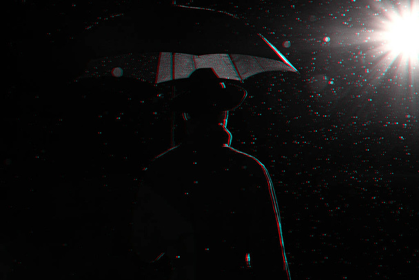 σκούρα σιλουέτα ενός άνδρα με αδιάβροχο και καπέλο κάτω από μια ομπρέλα στο δρόμο στη βροχή - Φωτογραφία, εικόνα