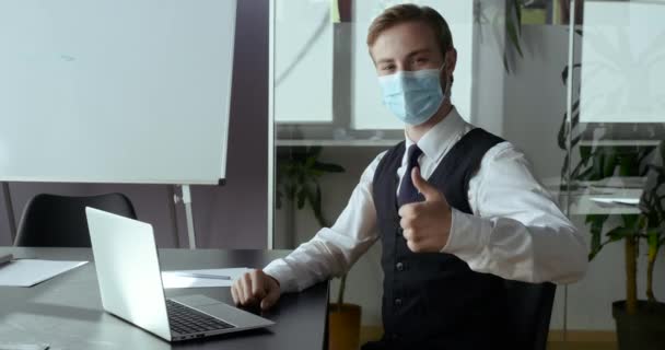 Young student guy successful business man wears protective medical mask on a male face, businessman sits in office shows sign of approval and support at camera with his hand, puts thumb up like symbol - Filmagem, Vídeo