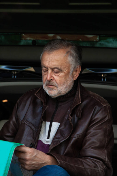 elderly man with a gray beard in a leather jacket folds a road map against the background of an open trunk of a ca - Zdjęcie, obraz