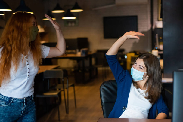 Co-workers in protective masks give a high five in the office. Women work during the coronavirus - Foto, Imagen