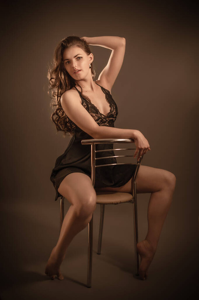 Portret van een jong mooi meisje dat haar lange donkere haar aanraakt poserend in ondergoed zittend op een stoel in de studio. - Foto, afbeelding