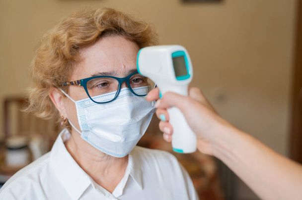 A doctor measures the temperature of an elderly woman wearing a mask with an infrared thermometer. house call medical check-up - Foto, immagini