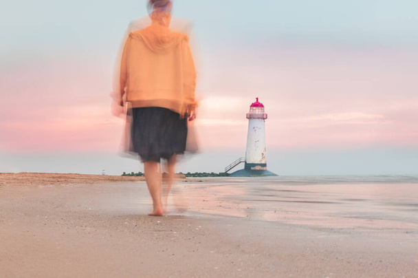 Movimiento borroso de la mujer en movimiento hacia el faro The Point of Ayr, también conocido como el faro Talacre, situado en la costa norte de Gales, Reino Unido, puesta de sol irreal con colores pastel - Foto, imagen