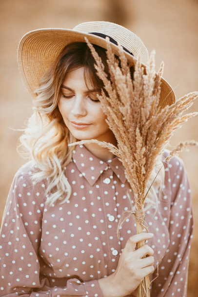 A young girl with long curly hair and a hat , in a brown dress with white polka dots, stands in a wheat field, against the background of the sunset - Φωτογραφία, εικόνα