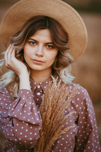 Uzun kıvırcık saçlı ve şapkalı, beyaz benekli kahverengi elbiseli genç bir kız, gün batımının arka planında, buğday tarlasında duruyor. - Fotoğraf, Görsel