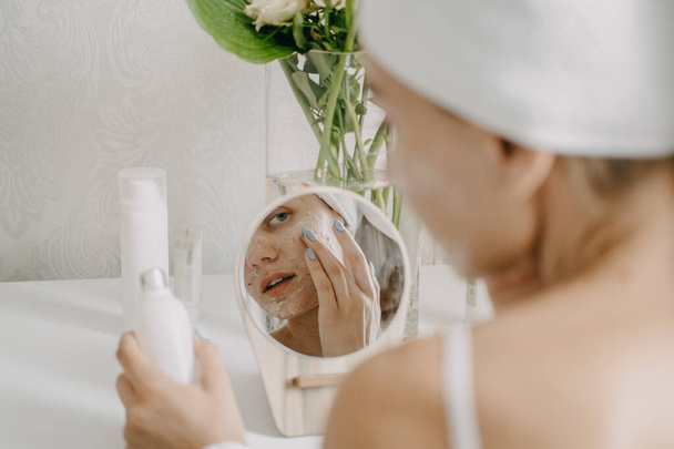 Jovem mulher em toalha branca refrigerando no quarto e fazendo máscara facial de barro perto do espelho. Menina fazendo tratamento de beleza e relaxar em casa. Cuidados com a pele da manhã rotina de beleza, autocuidado - Foto, Imagem