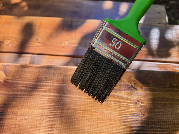 Εφαρμογή προστατευτικού βερνικιού σε ένα ξύλινο πάτωμα αίθριο.Ζωγραφική ξυλουργική έξω.hand χρήση πινέλο χρώμα σαφή λάκα στην επιφάνεια του ξύλου - Φωτογραφία, εικόνα