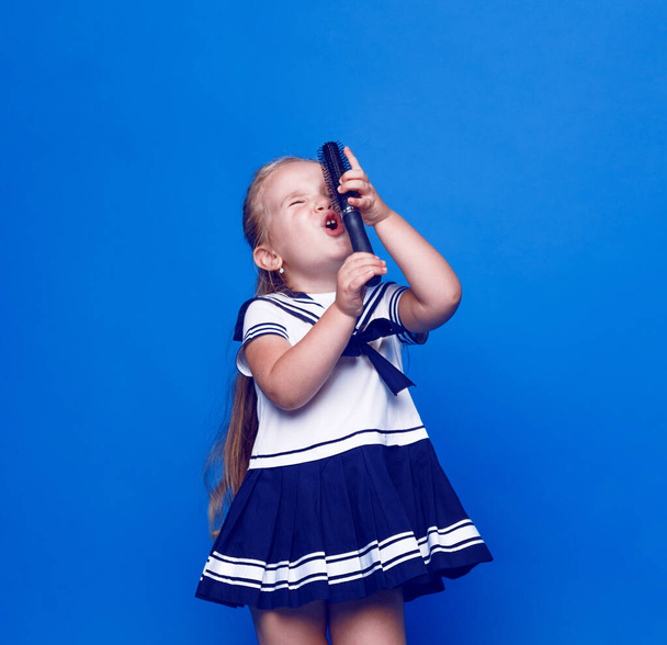 Αξιολάτρευτο κοριτσάκι με μπλε μάτια, ντυμένο με φόρεμα, κρατώντας μια χτένα αντί για μικρόφωνο σε μπλε φόντο. Θέση για κείμενο. - Φωτογραφία, εικόνα