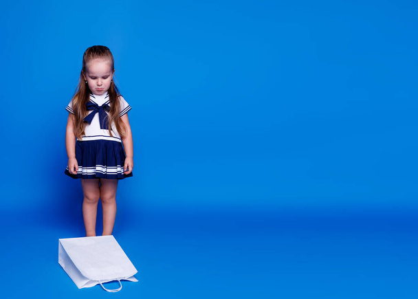 Несчастная маленькая симпатичная девочка в платье, глядя на белую сумку с покупками, сидящую на полу, на синем фоне. - Фото, изображение