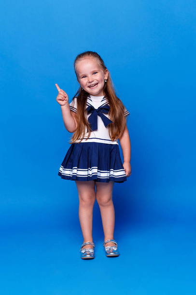 Schattig lachend meisje wijst met haar vinger omhoog, draagt een jurk, geïsoleerde blauwe achtergrond, studio opname. Verticaal overzicht. - Foto, afbeelding