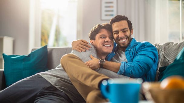 Portrait of a Cute Male Queer Couple at Home. They Sit on a Sofa and Look at the Camera. Partner Embraces His Lover from Behind. They are Happy and Smiling. Room Has Modern Interior. - Фото, зображення
