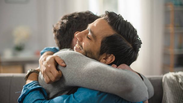 Cute Attractive Male Gay Couple Sit Together on a Sofa at Home. Boyfriends are Hugging and Embracing Each Other. They are Happy and Smiling. They are Casually Dressed and Room Has Modern Interior. - Valokuva, kuva