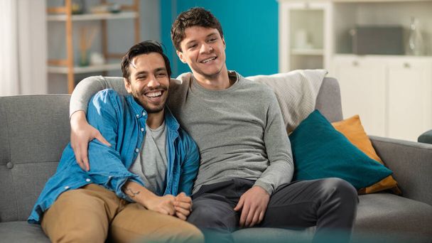 Portrait of a Cute Male Queer Couple at Home. They Sit on a Sofa. Partner Embraces His Lover from Behind. They are Happy and Smiling. Room Has Modern Interior. - Fotoğraf, Görsel