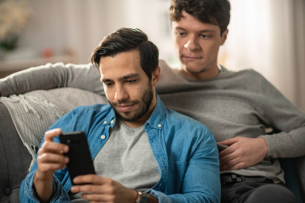 Cute Male Gay Couple Spend Time at Home. They are Lying Down on a Sofa and Use a Smartphone. They Browse Online. Partners Hand is Around His Lover. They Smile and Laugh. Room Has Modern Interior. - Foto, Imagem
