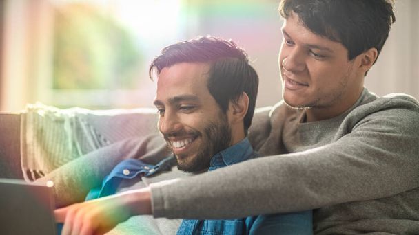 Male Queer Couple Spend Time at Home. They are Lying Down on Sofa and Use Laptop. They Browse Online. Partners Hand is Around His Lover and Pointing on Screen. Shot with Rainbow Lens Flare Effect. - Fotografie, Obrázek