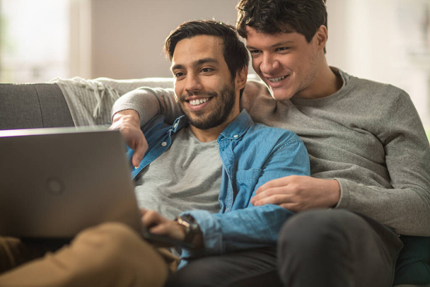 Sweet Male Queer Couple Spend Time at Home. They are Lying Down on a Sofa and Use the Laptop. They Browse Online. Partners Hand is Around His Lover. They Smile and Laugh. Room Has Modern Interior. - Fotografie, Obrázek