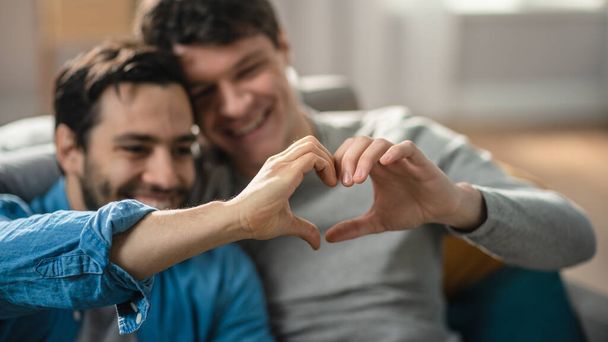 Portrait of a Cute Male Queer Couple at Home. They Sit on a Sofa and Making a Heart Symbol with Their Hands. Partner Has One Hand Behind His Lover. Room Has Modern Interior. - Foto, afbeelding