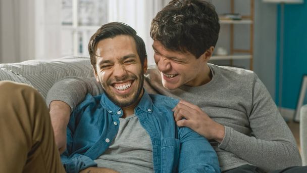 Portrait of a Cute Male Queer Couple at Home. They Sit on a Sofa and Joking. Partner Embraces His Lover from Behind. They are Happy and Smiling. Room Has Modern Interior. - Photo, image