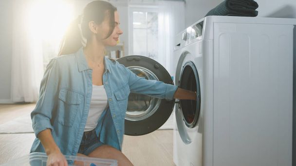 Beautiful Smiling Brunette Young Woman Sits in Front of a Washing Machine in Homely Jeans Clothes. She Loads the Washer with Dirty Laundry. Bright and Spacious Living Room with Modern Interior. - Fotoğraf, Görsel