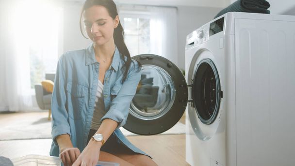 Beautiful Smiling Brunette Young Woman Sits in Front of a Washing Machine in Homely Jeans Clothes. She Loads the Washer with Dirty Laundry. Bright and Spacious Living Room with Modern Interior. - Fotoğraf, Görsel