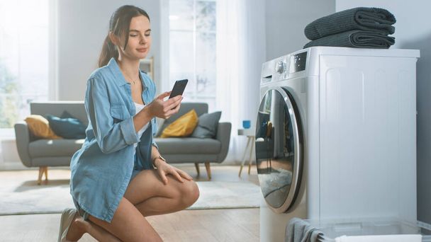 Beautiful Young Woman Sits on Her Knees Next to the Washing Machine. She Loaded the Washer with Dirty Laundry and Configured the Wash With Her Smartphone. Shot in Living Room with Modern Interior. - Foto, imagen