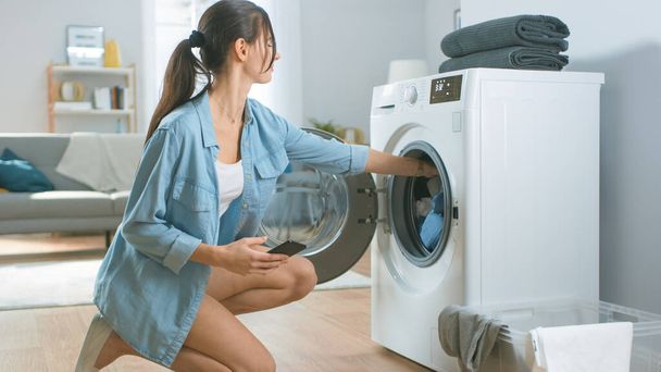 Beautiful Young Woman Sits on Her Knees Next to the Washing Machine. She Loaded the Washer with Dirty Laundry and Configured the Wash With Her Smartphone. Shot in Living Room with Modern Interior. - Foto, immagini