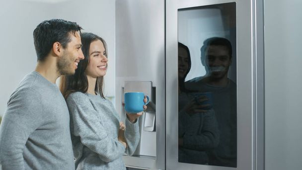 Beautiful Young Couple Drink Morning Coffee in the Kitchen. They Check the Weather Forecast and a To Do List on a Smart Fridge at Home. Apartment is Bright and Cozy. - Φωτογραφία, εικόνα
