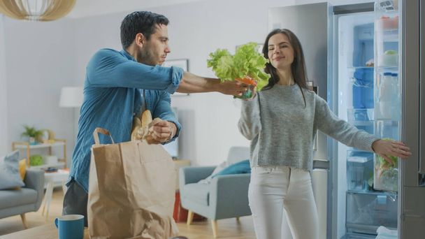 Beautiful Young Couple Come to the Kitchen with Fresh Groceries in Brown Paper Bag. Man is Handing Fresh Salad Greens and Oranges to the Girl Who Puts Them in the Fridge. - 写真・画像