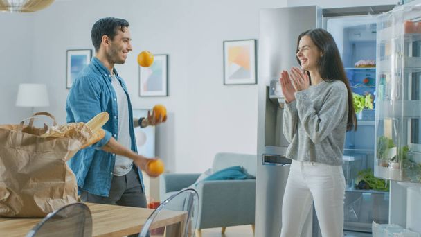 Beautiful Young Couple Have Fun in the Kitchen. Man is Juggling with Oranges. Girl is Clapping, Cheering for Him and They Laugh. Room Has a Modern Tech Fridge with Fresh Groceries. - Foto, afbeelding