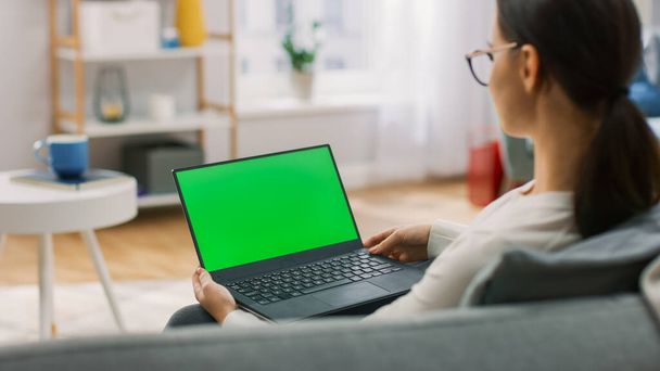 Young Woman at Home Sitting on a Couch Works on a Laptop Computer with Green Mock-up Screen. Girl Using Computer, Browsing through Internet, Watching Content, Chatting in Social Networks with Friends. - 写真・画像