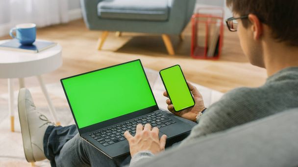 Young Man at Home Works on a Laptop Computer with Green Mock-up Screen, while Holding Smartphone with Chroma Key Display. Hes Sitting On a Couch in His Cozy Living Room. Over the Shoulder Camera Shot - Фото, изображение