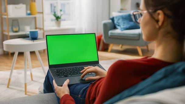 Young Woman at Home Works on a Laptop Computer with Green Mock-up Screen. Shes Sitting On a Couch in His Cozy Living Room. Over the Shoulder Shot - Foto, afbeelding