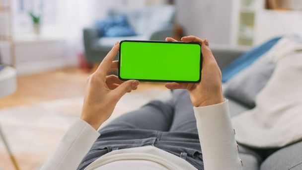 Young Woman at Home Laying on a Couch using with Green Mock-up Screen Smartphone in Horizontal Landscape Mode. Girl Using Mobile Phone, Browsing Internet, Watching Content, Videos, Blogs. POV. - Photo, image