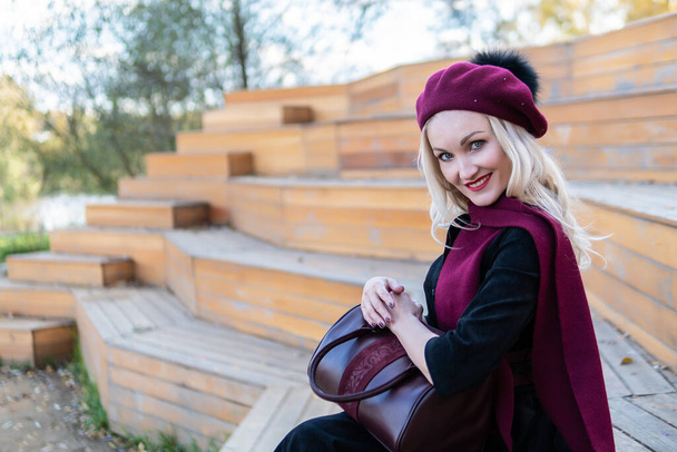 A smiling woman sits on a summer theater bench, made of wood in a burgundy coat and biret, an adult looks at the camera, in the fall against a background of blue clouds. - Фото, изображение