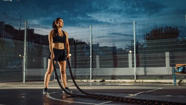 Beautiful Energetic Fitness Girl Doing Exercises with Battle Ropes. She is Doing a Workout in a Fenced Outdoor Basketball Court. Evening After Rain in a Residential Neighborhood Area. - Zdjęcie, obraz