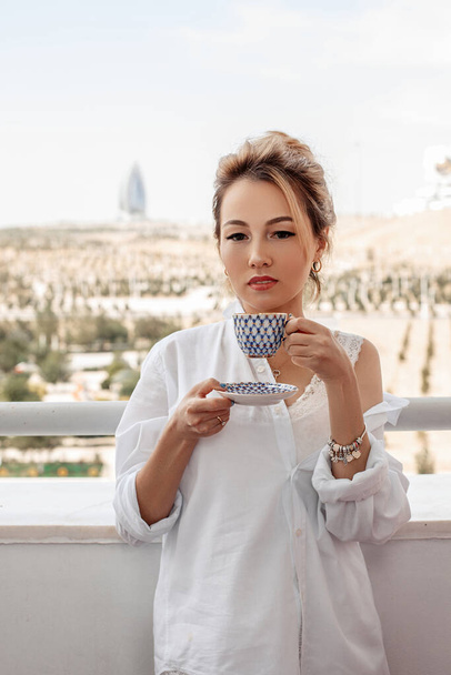 юна красива азіатська дівчина в бежевих мереживних шортах, біла сорочка п'є каву стоячи на балконі. вибірковий фокус. маленька зона фокусу. - Фото, зображення