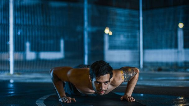 Strong Muscular Fit Shirtless Young Man is Doing Push Up Exercises. He is Doing a Workout in a Fenced Outdoor Basketball Court. Night After Rain in a Residential Neighborhood Area. - 写真・画像