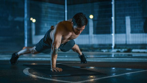 Strong Muscular Fit Shirtless Young Man is Doing One-Hand Push Up Exercises. He is Doing a Workout in a Fenced Outdoor Basketball Court. Night After Rain in a Residential Neighborhood Area. - Foto, Imagen