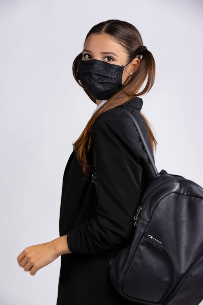 A young female student wearing a black face mask to prevent infection with the corona virus, wears a black jacket and a backpack. Photo on gray background. High quality photo - Photo, Image