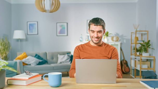 Portrait of Handsome Smiling Man Working on Laptop, Sitting at His Wooden Desk at Home. Man Browsing Through Internet, Working on Notebook from His Living Room. - Photo, Image