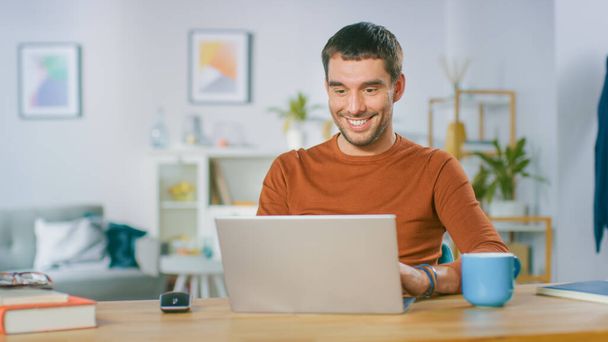 Portrait of Handsome Smiling Man Working on Laptop, Sitting at His Wooden Desk at Home. Man Browsing Through Internet, Working on Notebook from His Living Room. - Foto, afbeelding